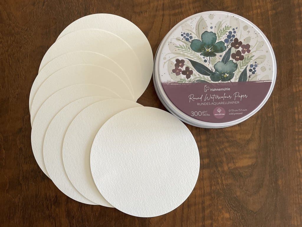 A set of round off-white watercolour paper circles fanned out. Hahnemuhle round tin container, 140lbs, 5 inches.