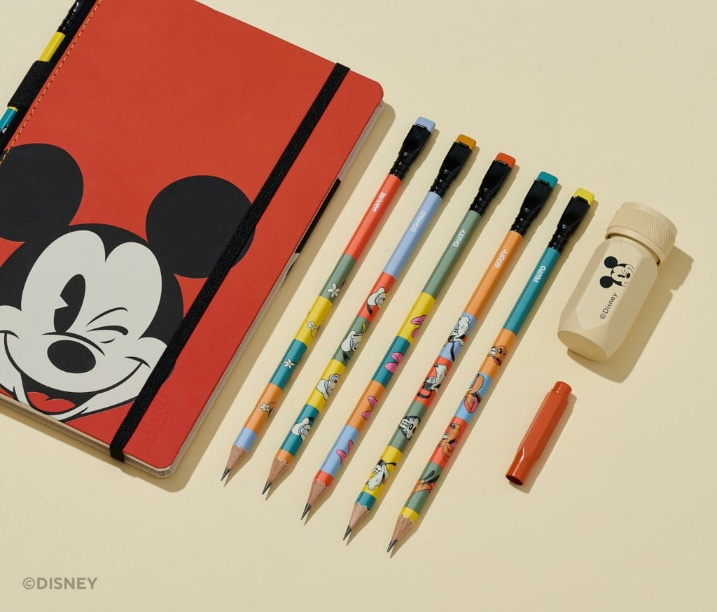 A Disney stationery set arranged on a warm cream background, including a red notebook with a Mickey Mouse face, five colorful pencils, a small orange pen cap, and a beige eraser with a Mickey Mouse head print.