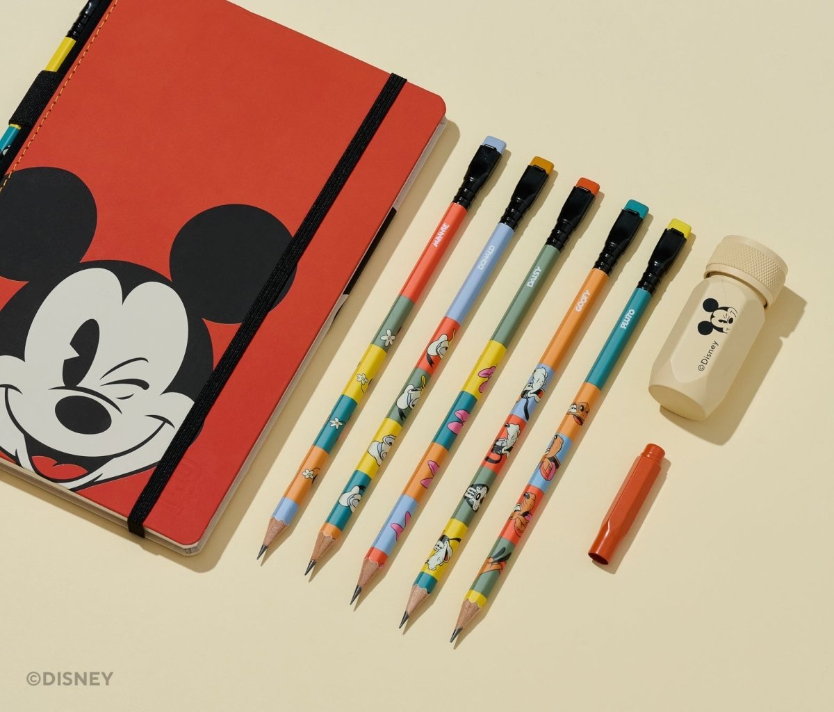 A Disney stationery set arranged on a warm cream background, including a red notebook with a Mickey Mouse face, five colorful pencils, a small orange pen cap, and a beige eraser with a Mickey Mouse head print.